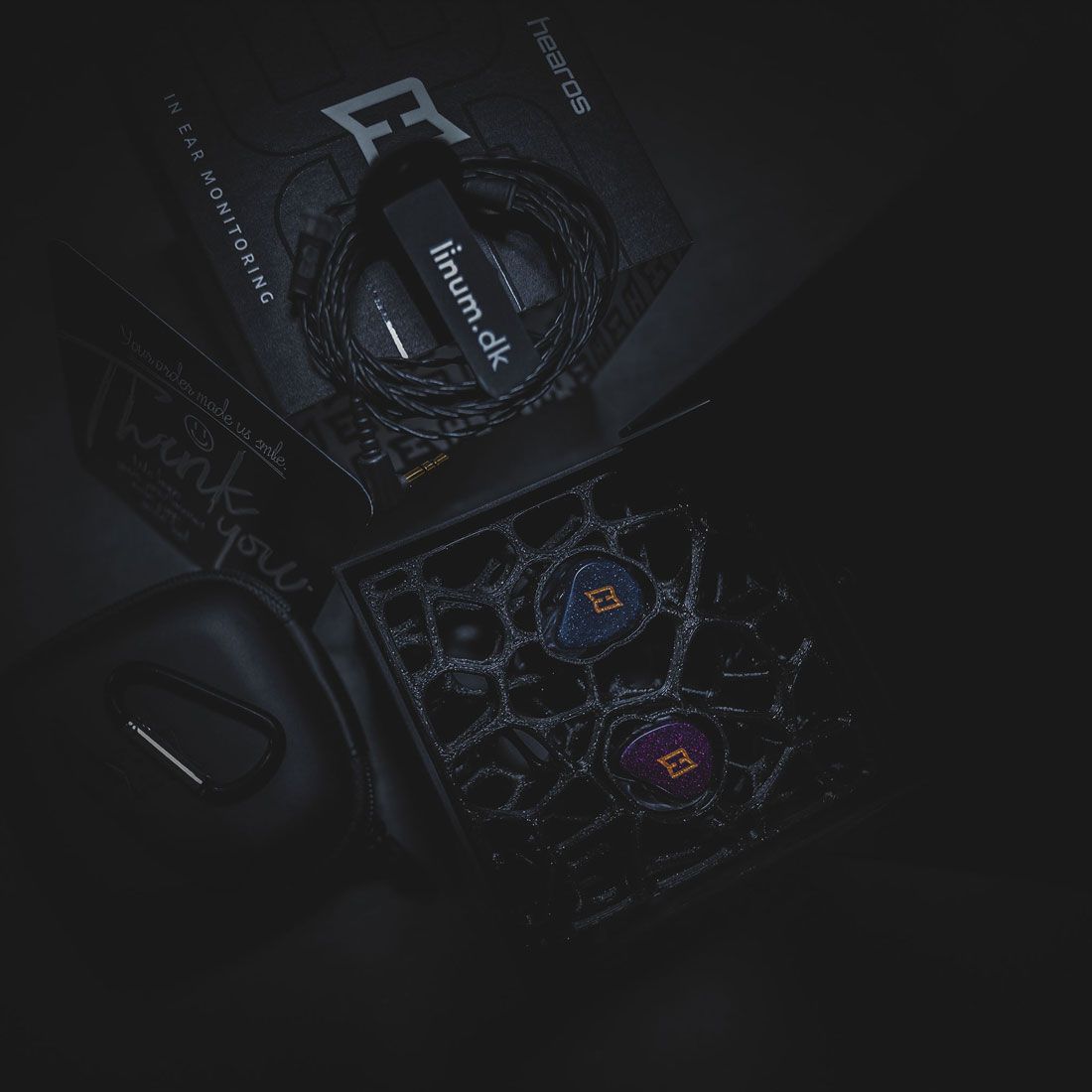 Premium product presentation of the hearos Customfit in-ear monitor package. The image shows customfit in-ear monitors in a presentation box, a Linum SuperBaX cable, a hearos carrying pouch, and branded packaging in a dark, high-end setting.