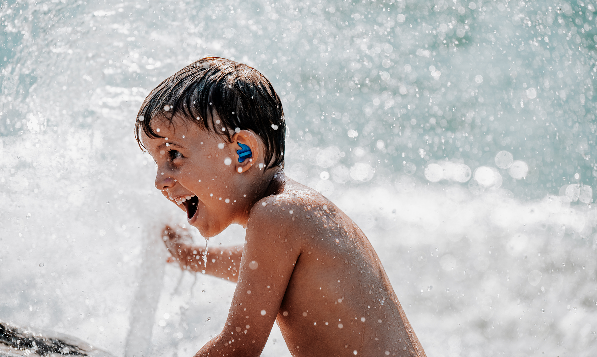 A joyful child with bachmaier® water earplugs plays under a spray of water, smiling widely, with droplets glistening in the sunlight.