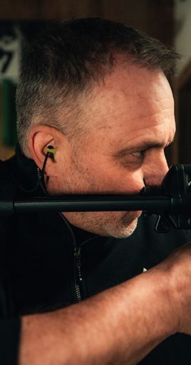 Person wearing custom hearing protection while aiming with an optical device in a tactical or security-related environment. Custom-fit hearing protection helps reduce exposure to loud impulse noise while maintaining comfort and situational awareness, making it ideal for security personnel, law enforcement training, and professional operational environments.