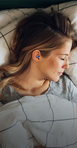 Person sleeping in bed while wearing custom hearing protection earplugs. Custom-fit hearing protection helps reduce disturbing noise during sleep, supporting a quieter and more comfortable sleeping environment at home, while traveling, or in noisy surroundings.