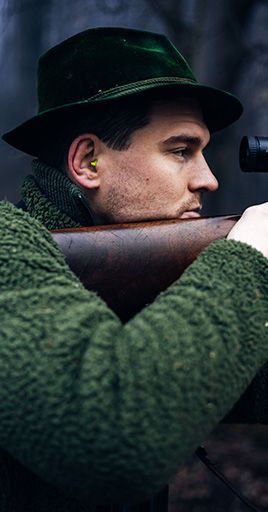 Hunter wearing custom hearing protection while aiming with a rifle in an outdoor environment. Custom-fit hearing protection helps reduce exposure to loud impulse noise during hunting activities while maintaining comfort and situational awareness in the field.